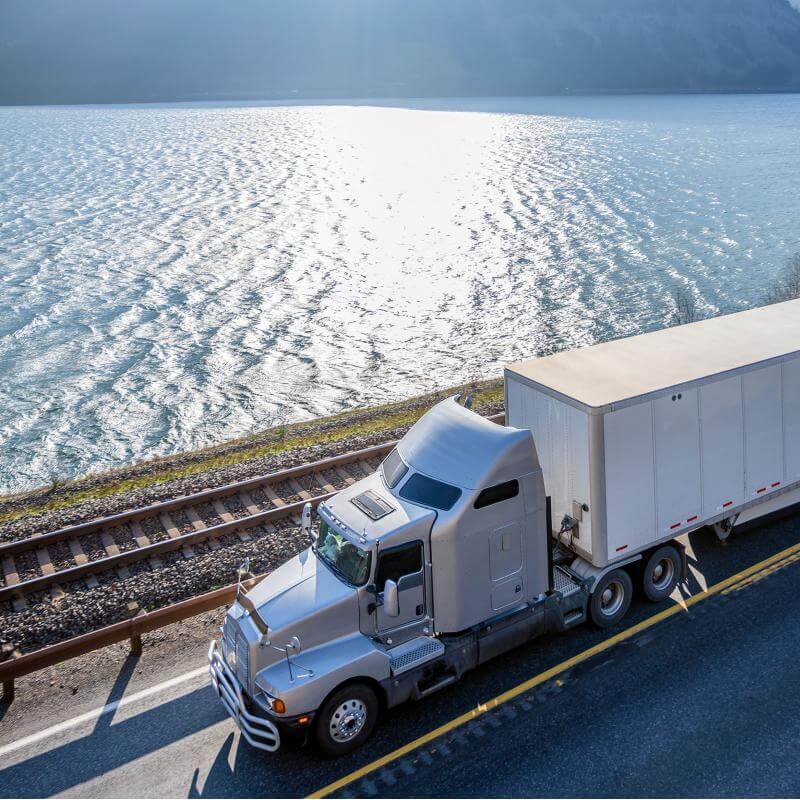 White commercial truck driving next to a rail track and water